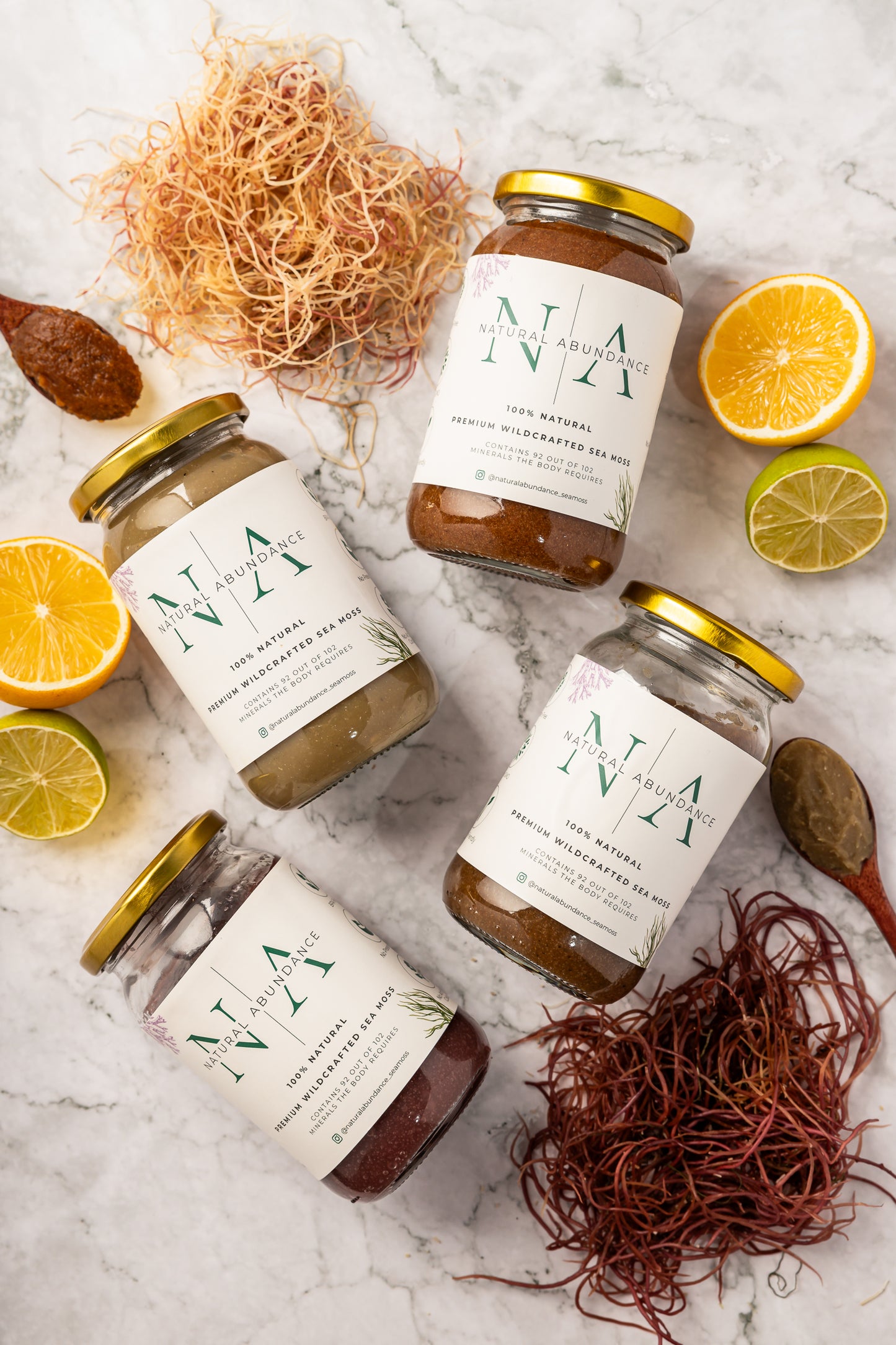 Four jars of Natural Abundance Premium Wildcrafted Sea Moss arranged on a marble surface with sea moss strands, lemon, and lime slices.