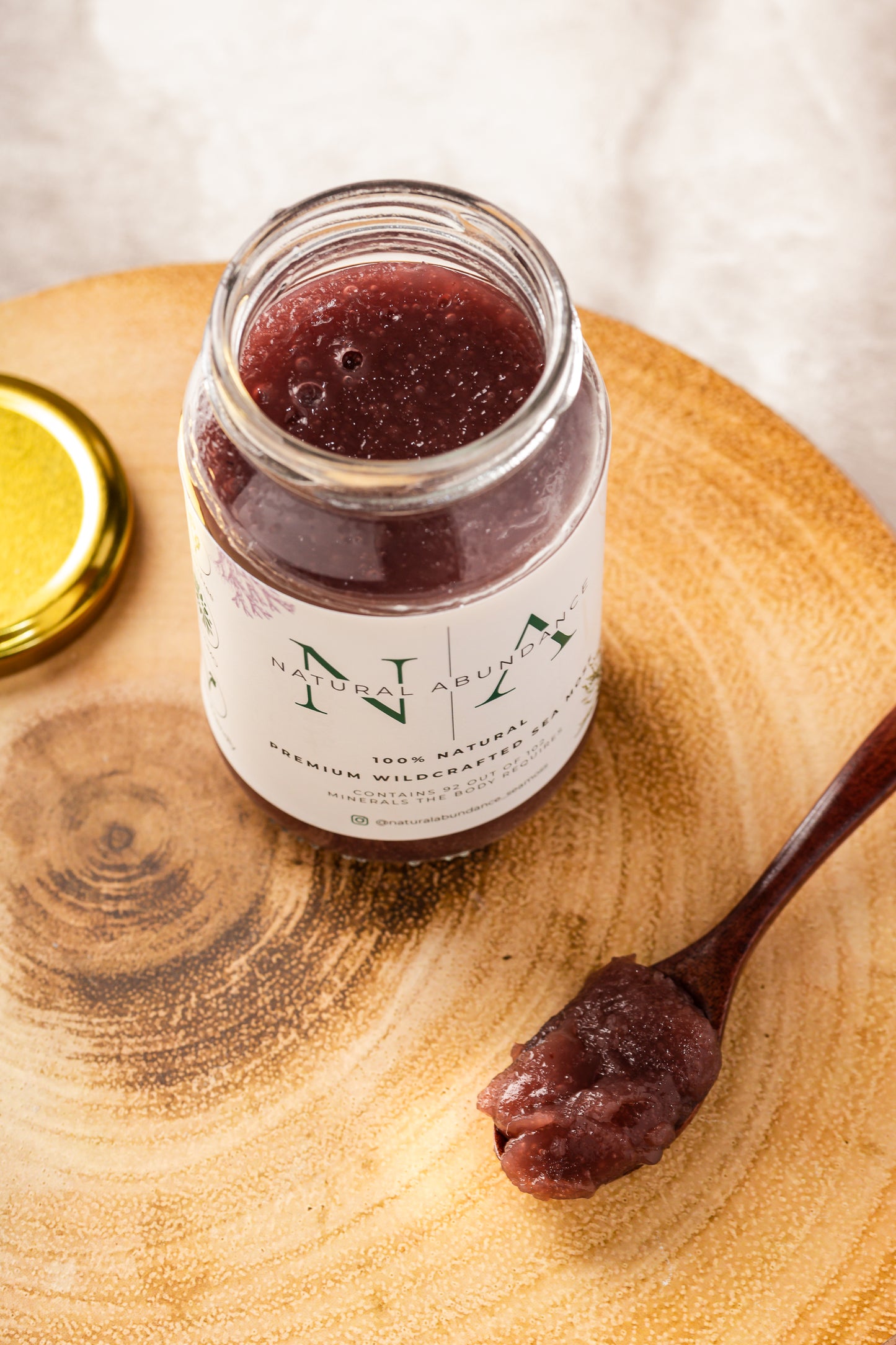 Open jar of Natural Abundance Premium Wildcrafted Sea Moss Gel with a wooden spoonful of purple sea moss on a wooden surface.